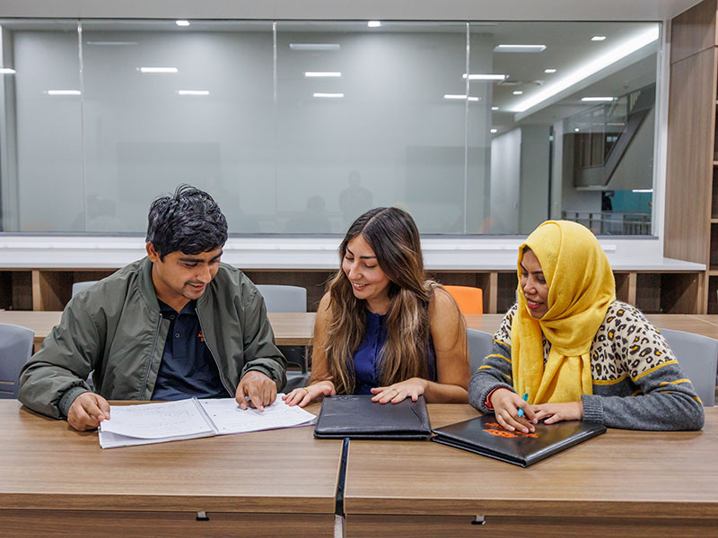 Three students sitting at their desks, looking together at a single notebook.