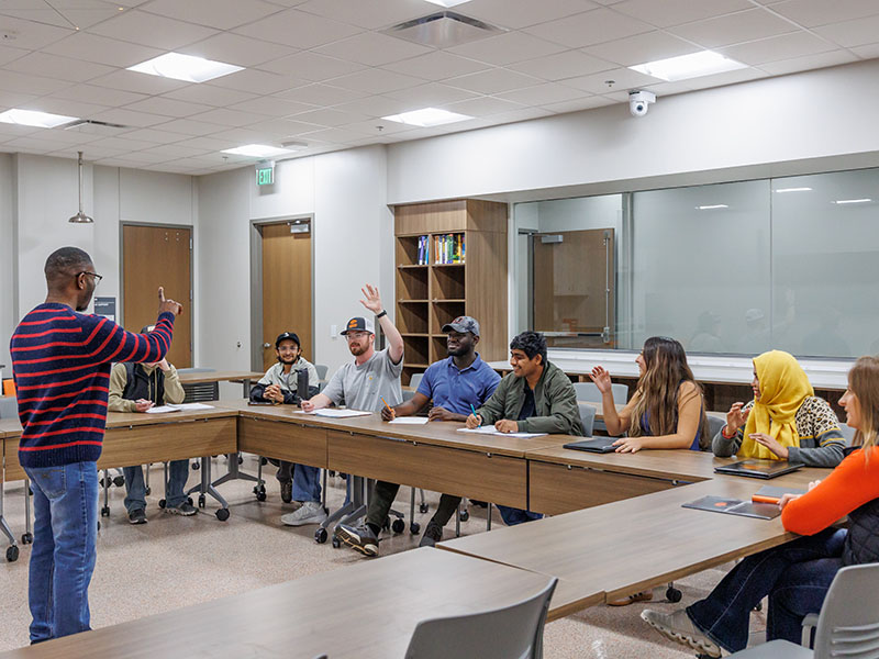 An instructor standing in a classroom with a few students seated in a U-shaped arrangement around him.
