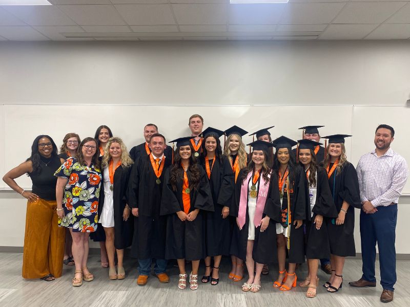 Spring 2025 Ag Leadership graduates standing in their graduation regalia posed for a photo. Accompanying them are three ag lead faculty dressed in professional clothing. All are smiling while standing in a classroom, in front of a white board.