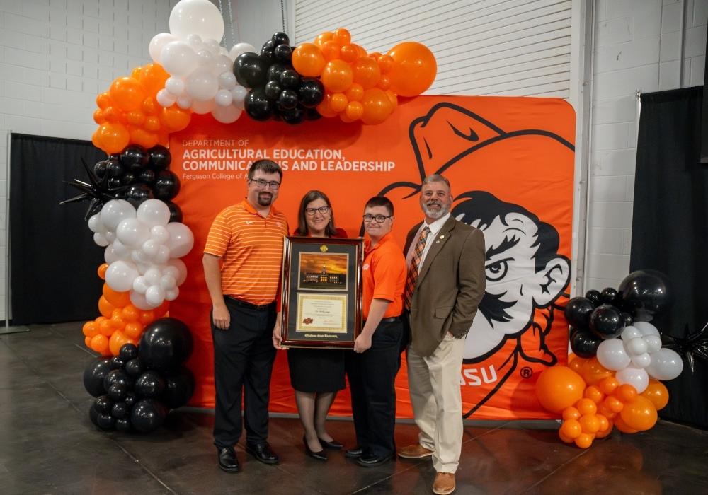 living-her-dream-1000x700px.jpg Legg is standing with her two sons and husband while holding a large award plaque. All four of them are dressed in business professional clothing. They are standing in front of an AECL department backdrop with a large balloon arch.