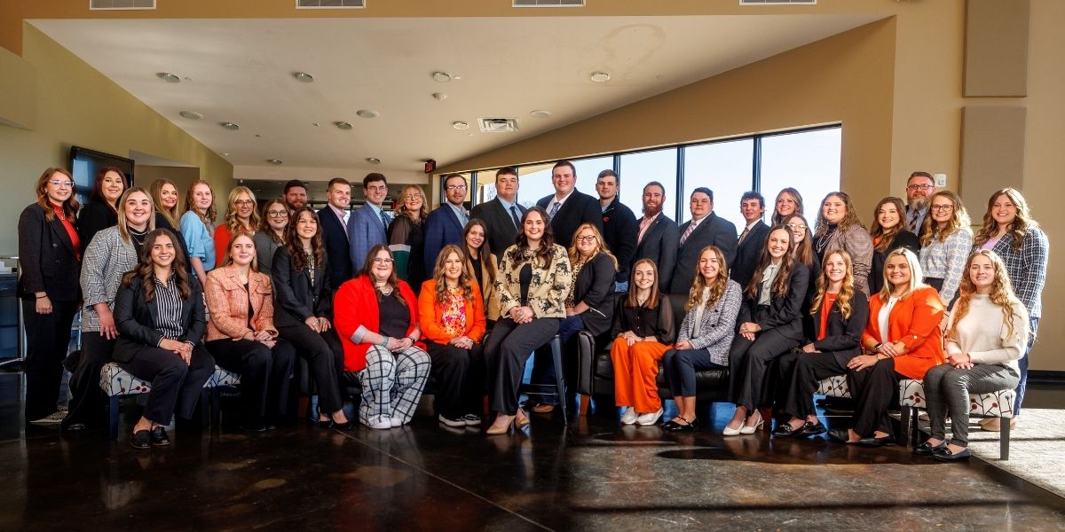 Spring 2025 student teachers pose for a group photo. All wearing professional dress clothing, some are standing some are sitting in chairs in front of the standing students.