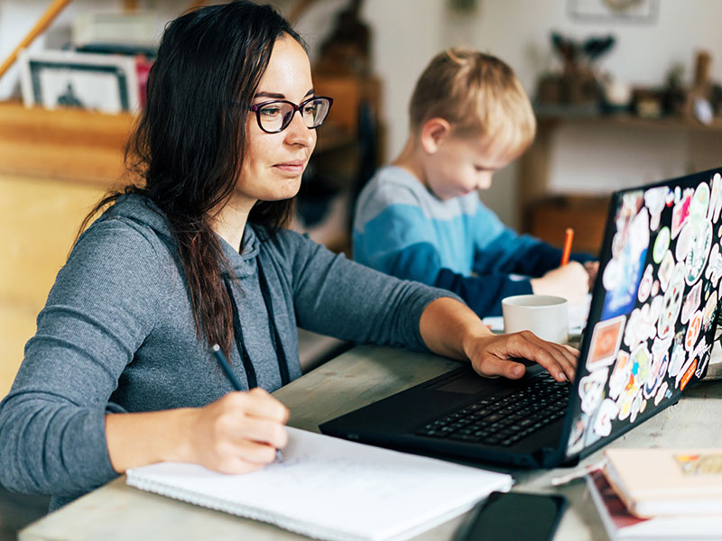 Woman working on her computer while her son plays at a kitchen table.