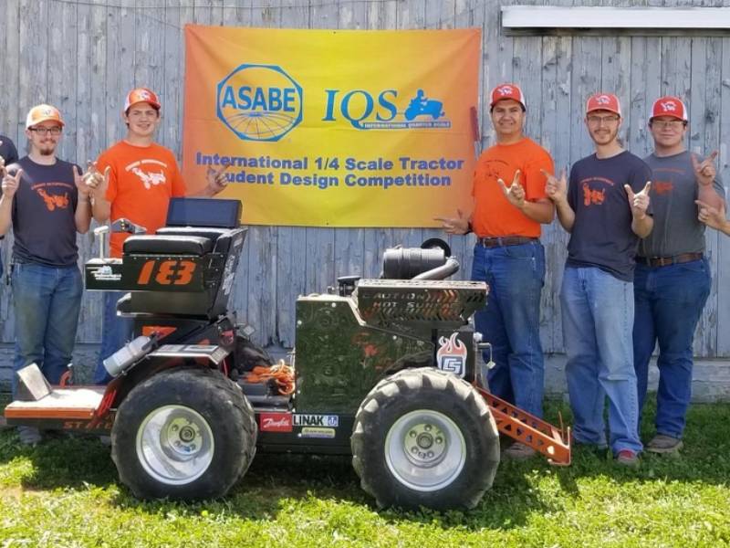 7 people standing near a tractor in front of a barn.