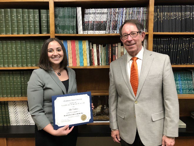 Ally Valdez is accepting her award from Jim Ansley in front of a book shelf.