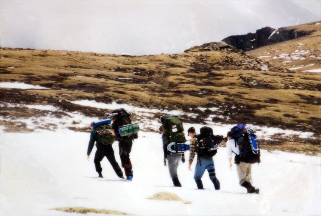 A group of five students carrying hiking gear as they tread through bright white snow near a hill.
