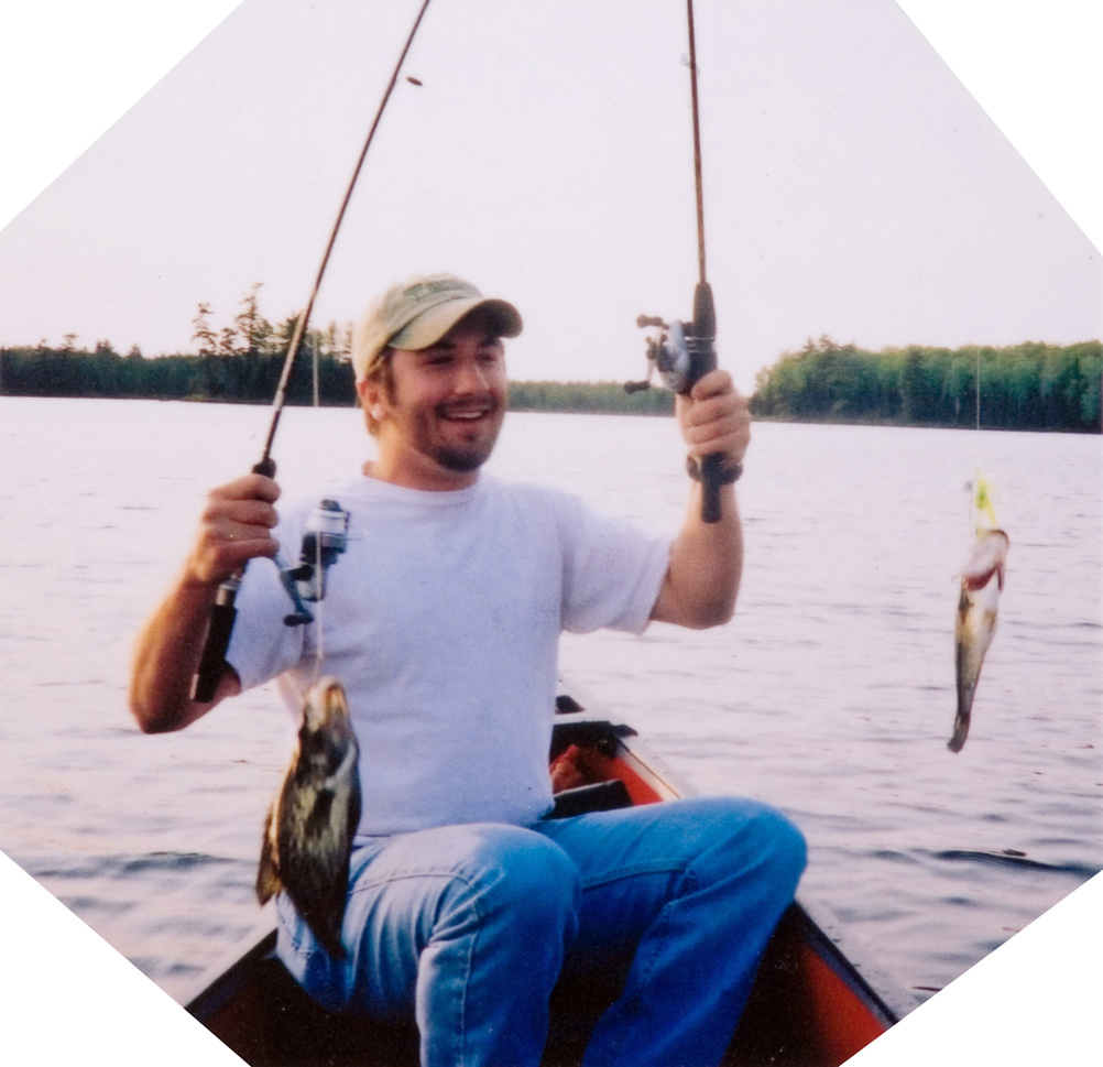 A student posing with two caught fish on poles inside of a boat on a lake.
