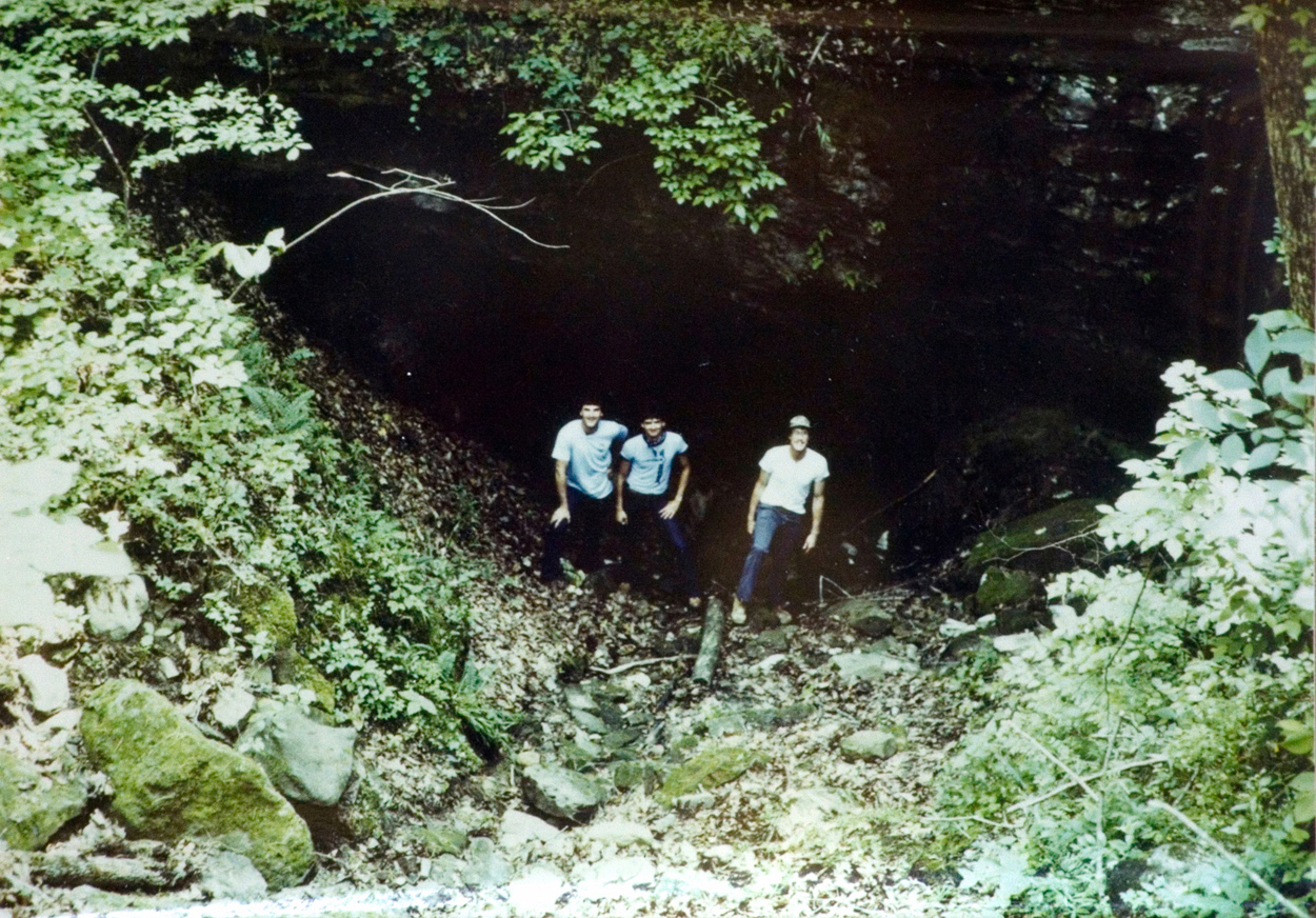 Three students pose in front of a cave entrance in the woods.