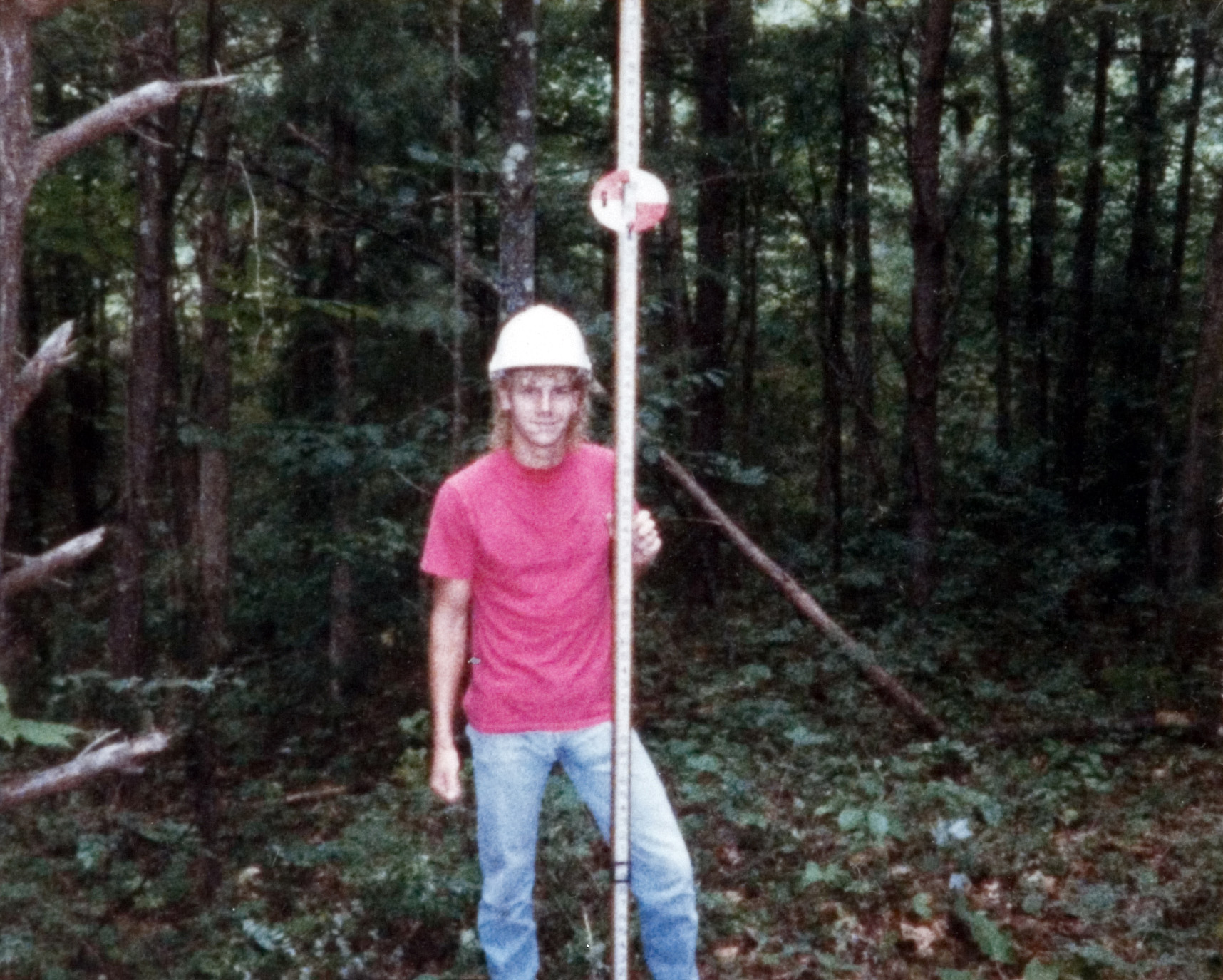 A student in a white hardhat pictured posing outside with a measuring tool.