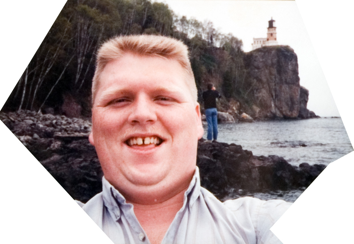 A male student takes a selfie near a cliff, lighthouse and shore.