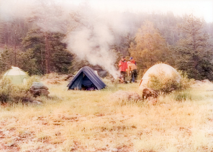 A field view of the student campsite at the base of a hill with a blue tent and a smokey fire going.