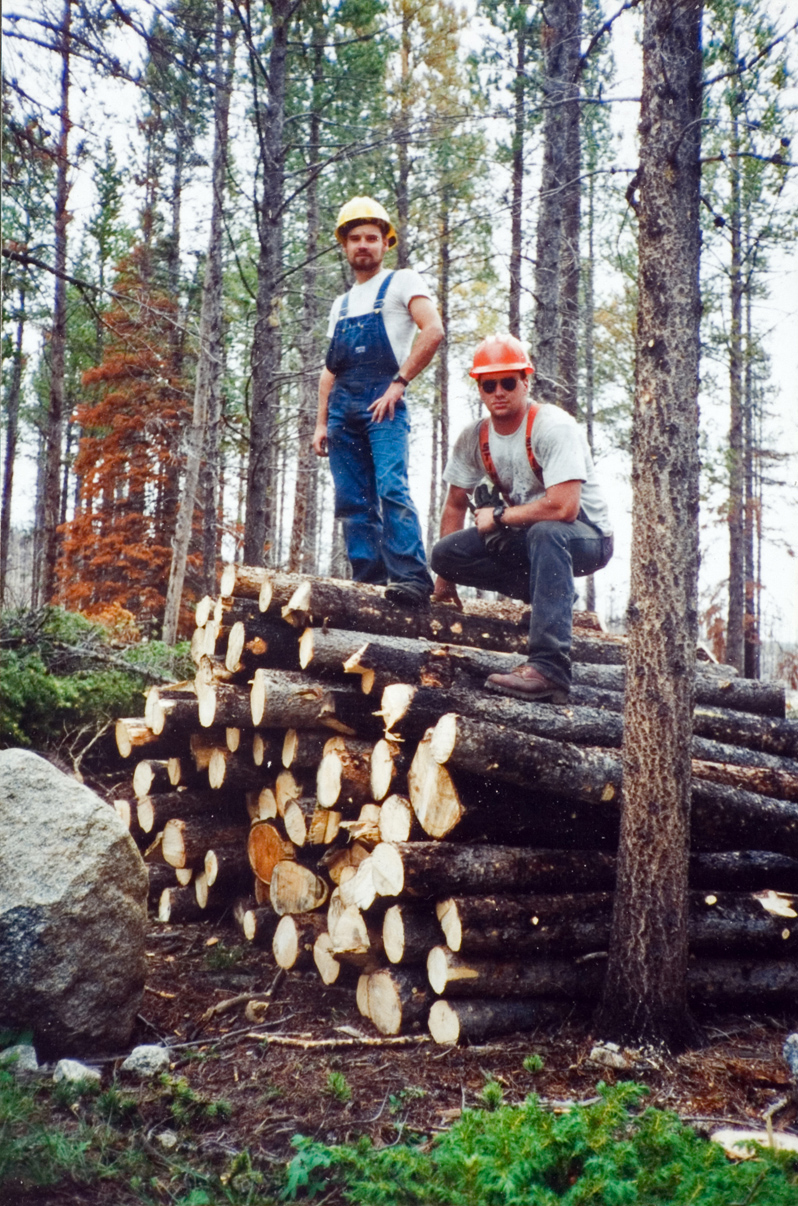 Two students in overalls and hardhats pose for the camera on top of a stack of lumber.
