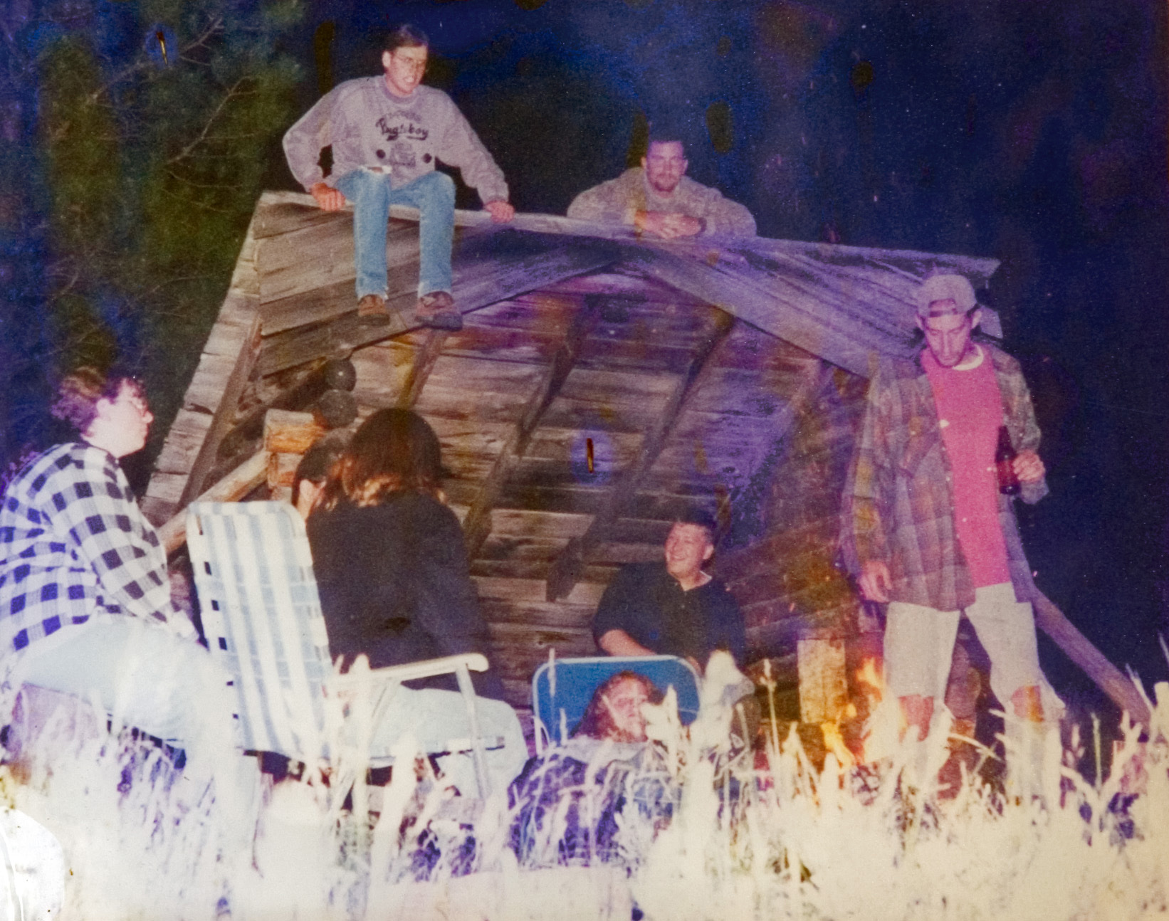 Six students gathered under a wooden covering as they converse with one another at a campsite.