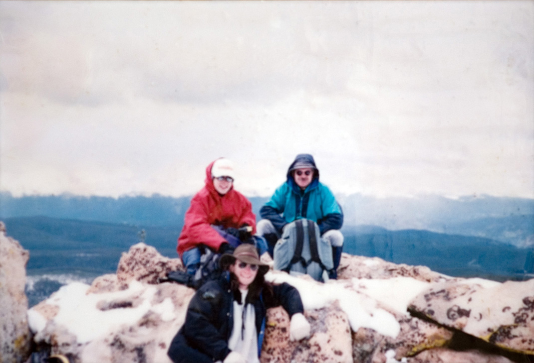 Three students in jackets sitting and standing alongside a snowy hilltop.