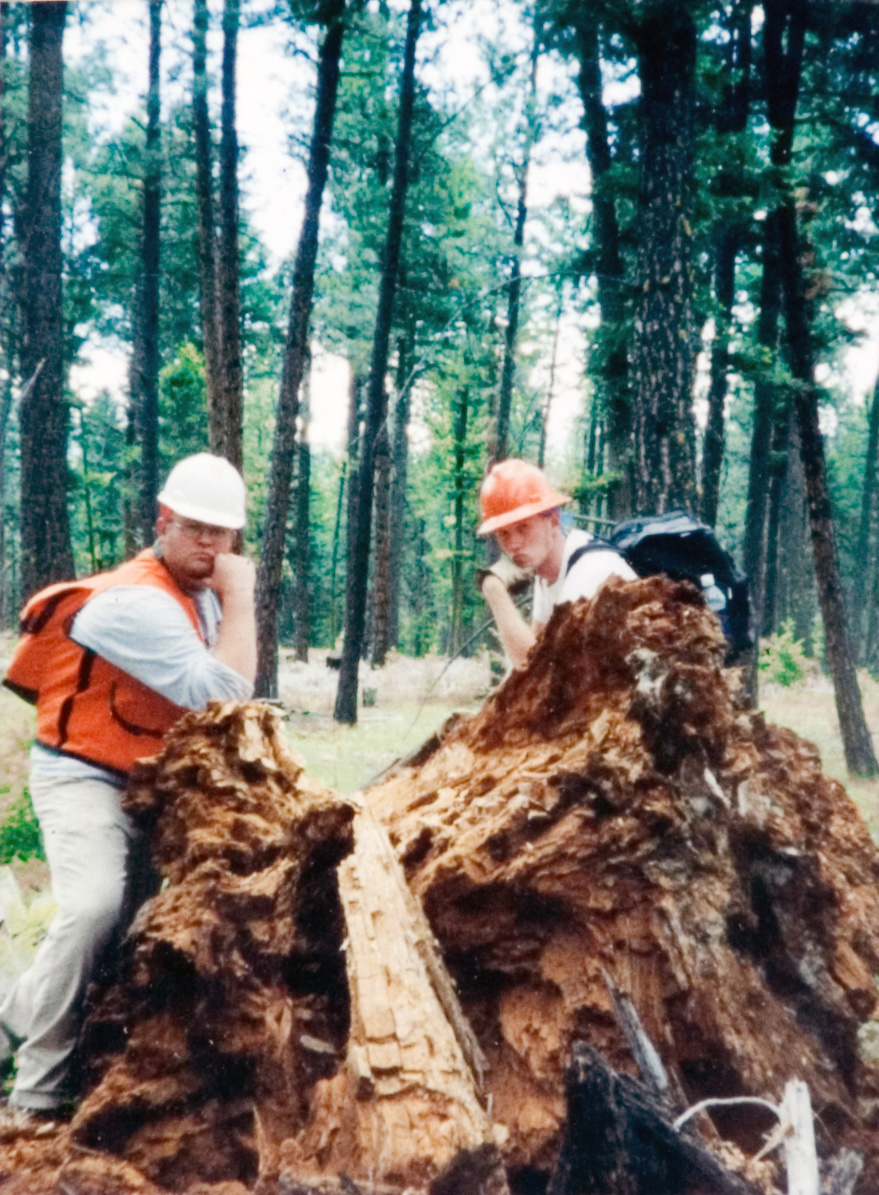 Two students in hardhats posing behind a large uprooted tree.