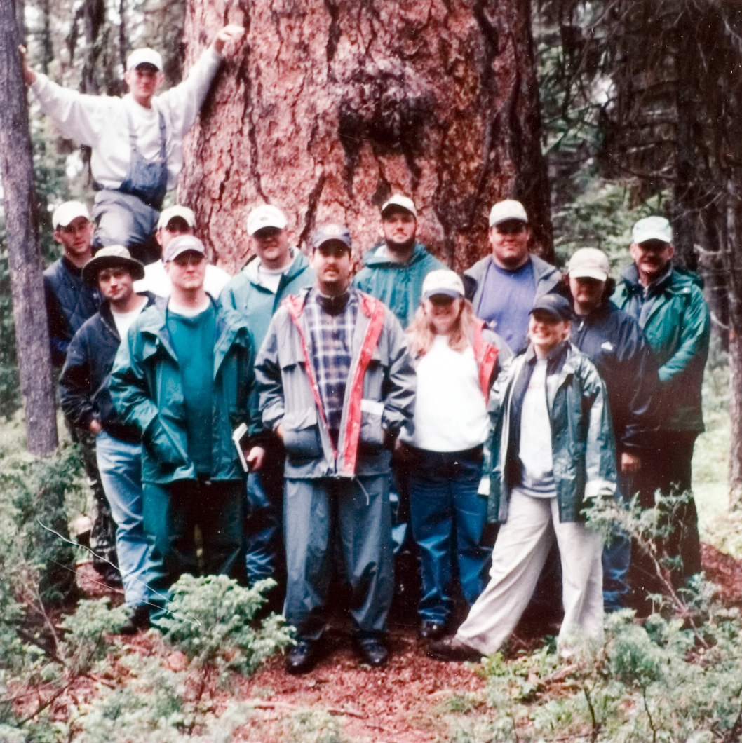 A group of 13 students in various attire as they stand in front of a large tree outside.