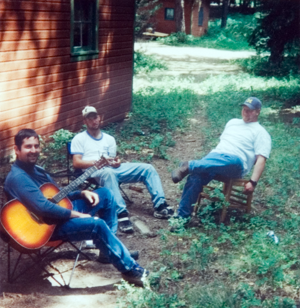 Three students sitting outside on the grass as one plays the guitar.