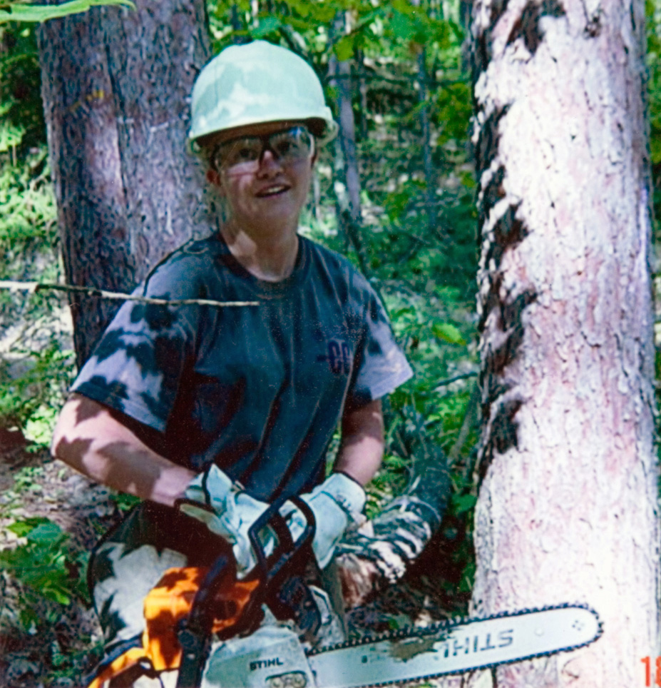 An individual in a hard hat posing with a chainsaw, preparing to cut a tree that is outside.