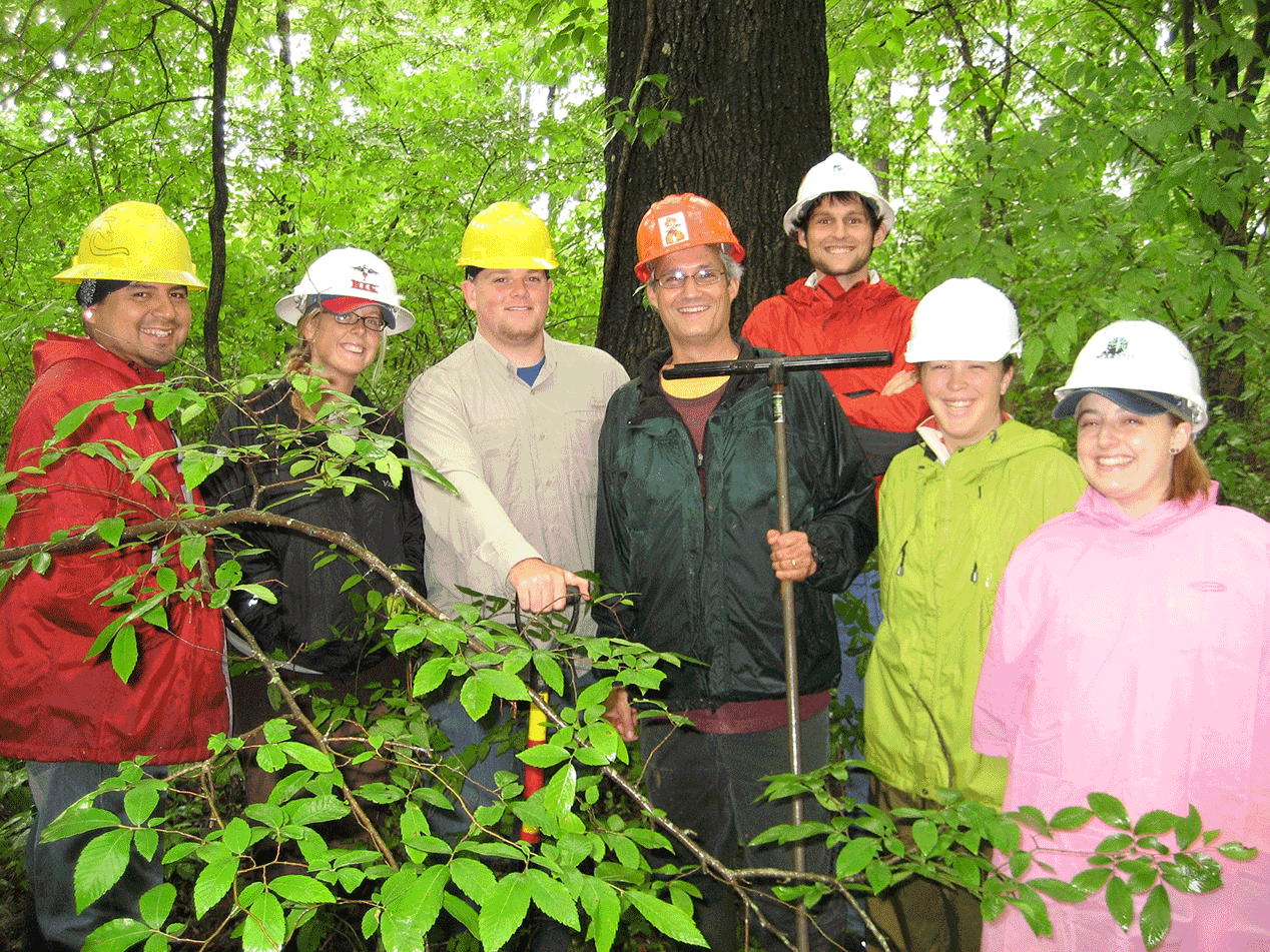 A group of people standing in hardhads in the woods smiling for the camera. 