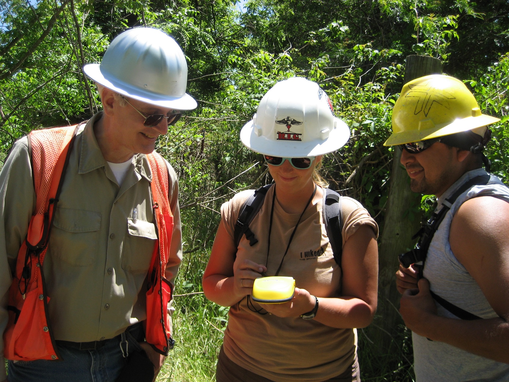 Three people wearing hardhats and taking notes outside. 
