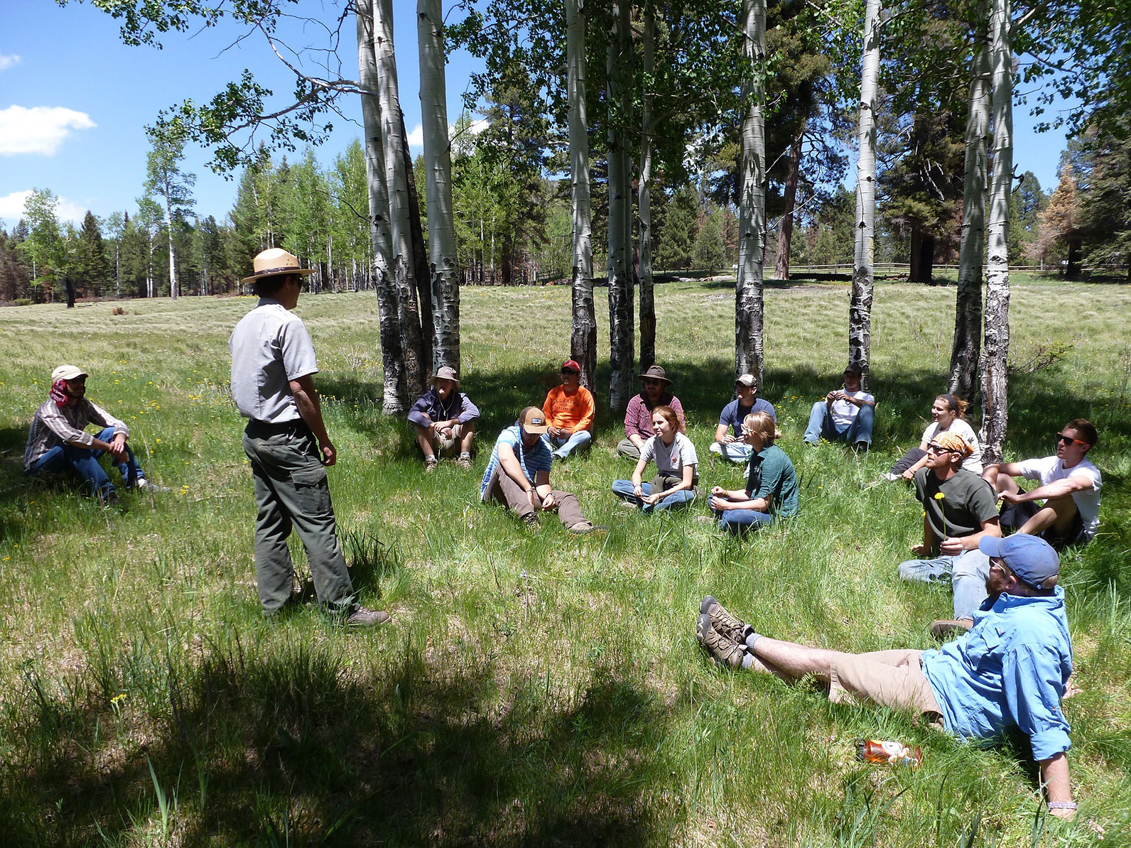 A group of participants sitting in the grass in a valley receiving directions from an instructor standing in the middle. 