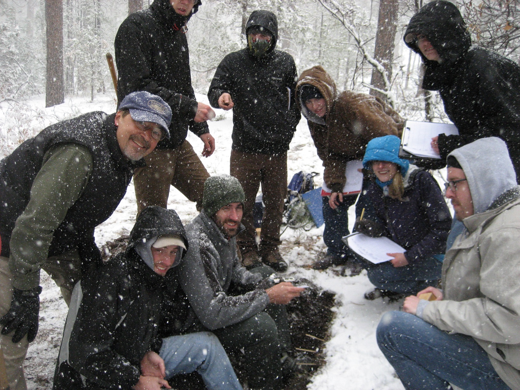 Nine individuals in a circle, in the snow, with clipboards, collecting information in the woods. 