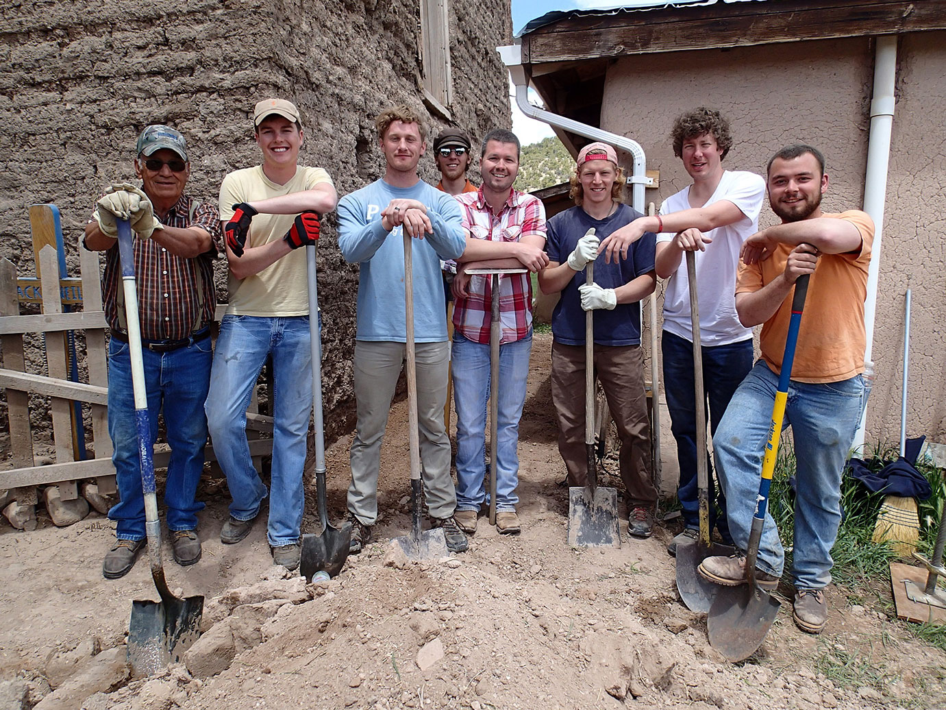 A group of eight people standing with shovels at the back of two buildings.