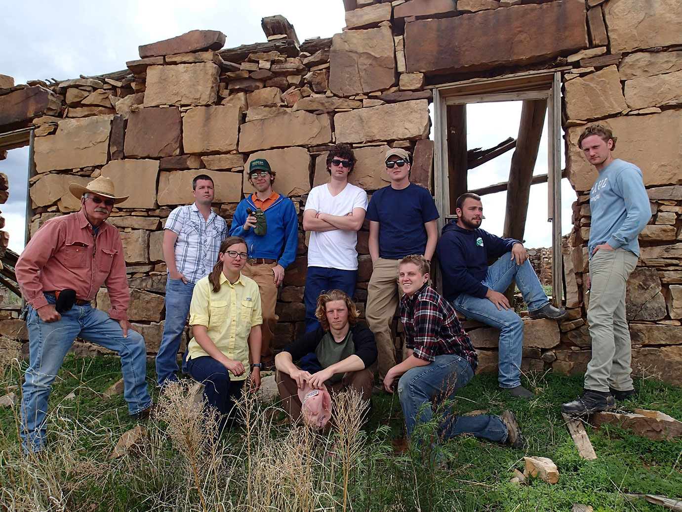 A group of ten people in front of a stone wall that is falling down.