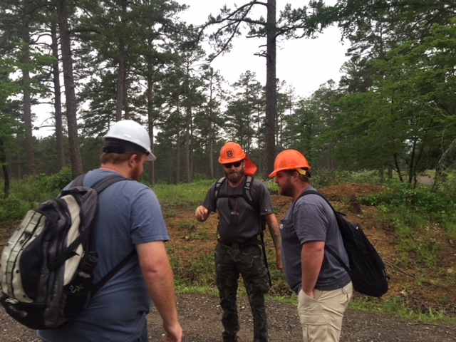 Three people in hardhats standing in a circle talking in a forest. 