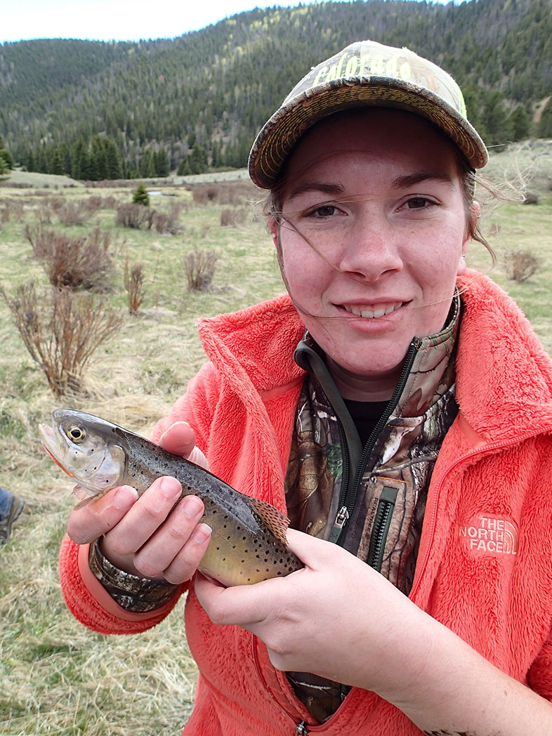 A girl in an orange jacket holding a fish and smiling at the camera. 