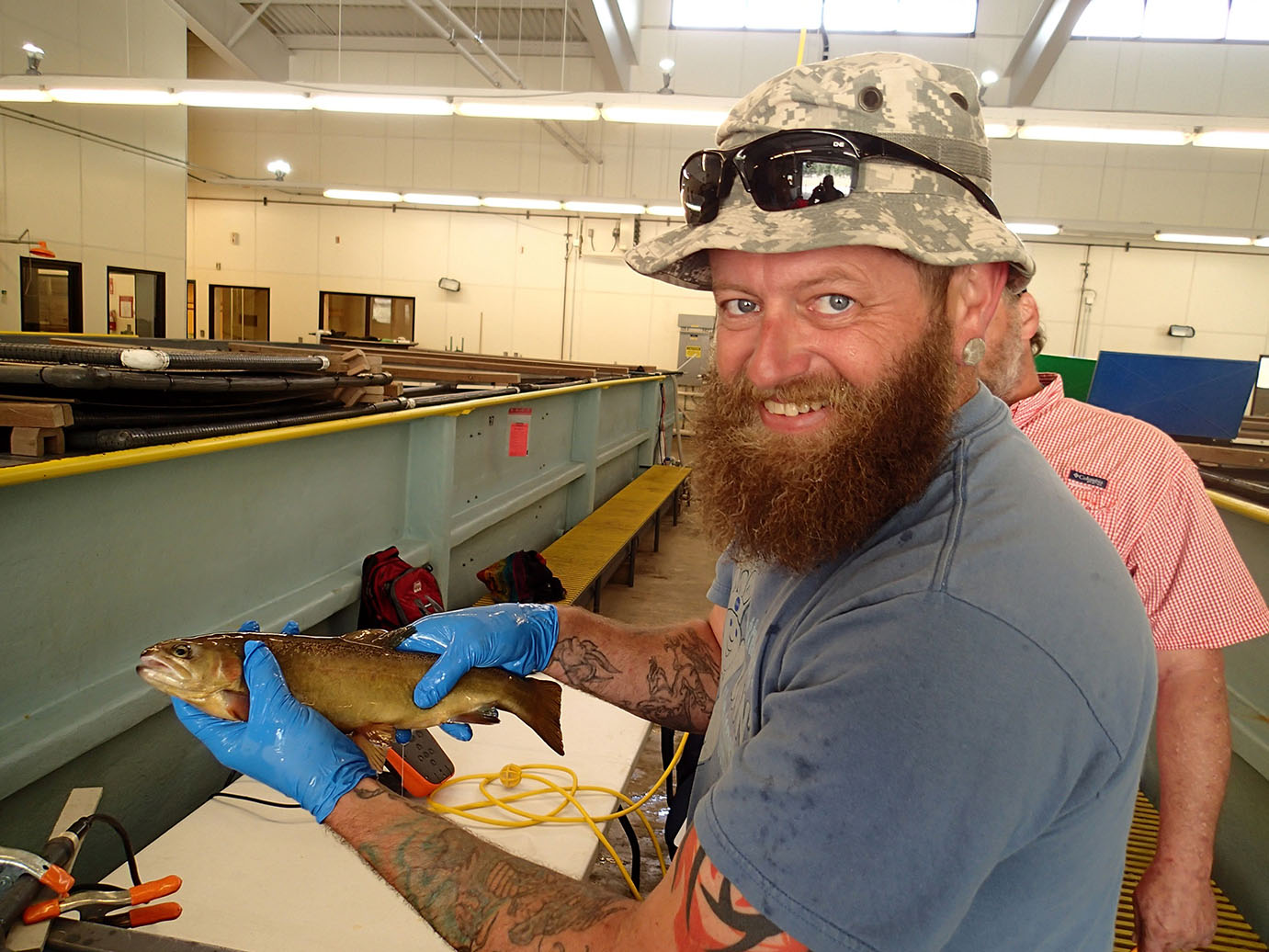 A man in a beard and a camouflage hat holding a fish inside a lab. 