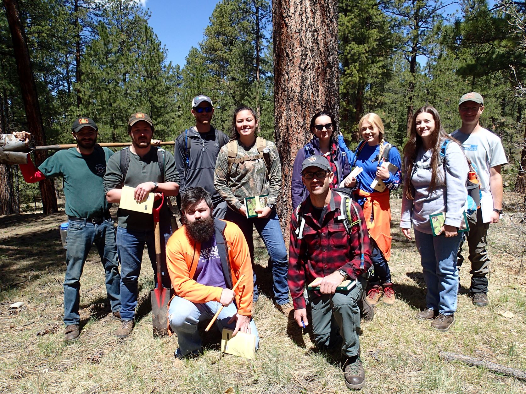 A co-ed group of people standing together in front of a tree in the woods. 