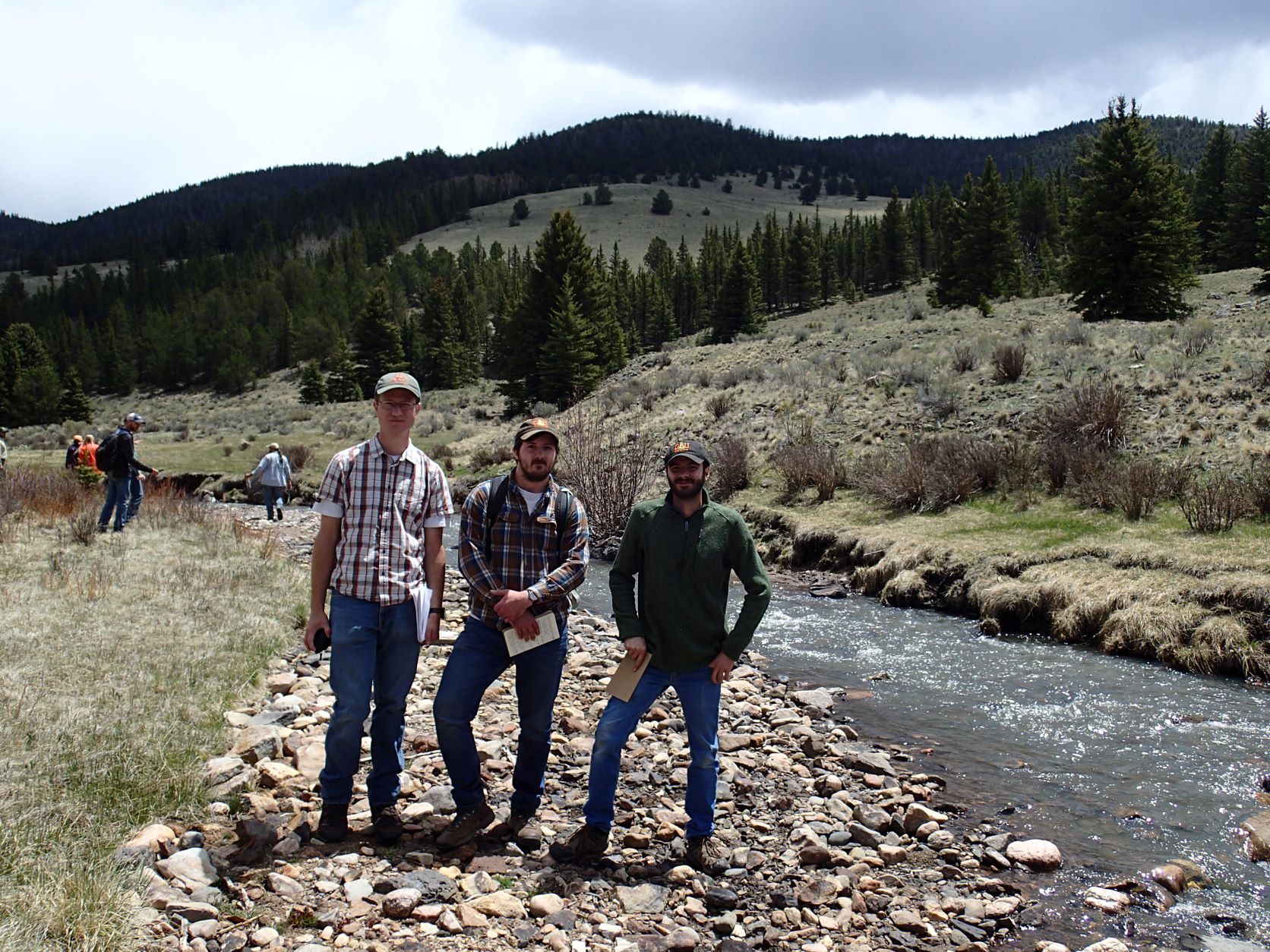 Three guys standing next to a small river running through a valley.