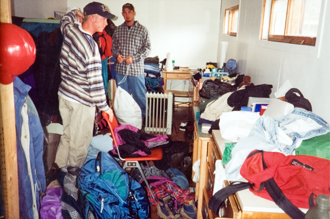 Two students in hats look around a messy room with wooden furniture.