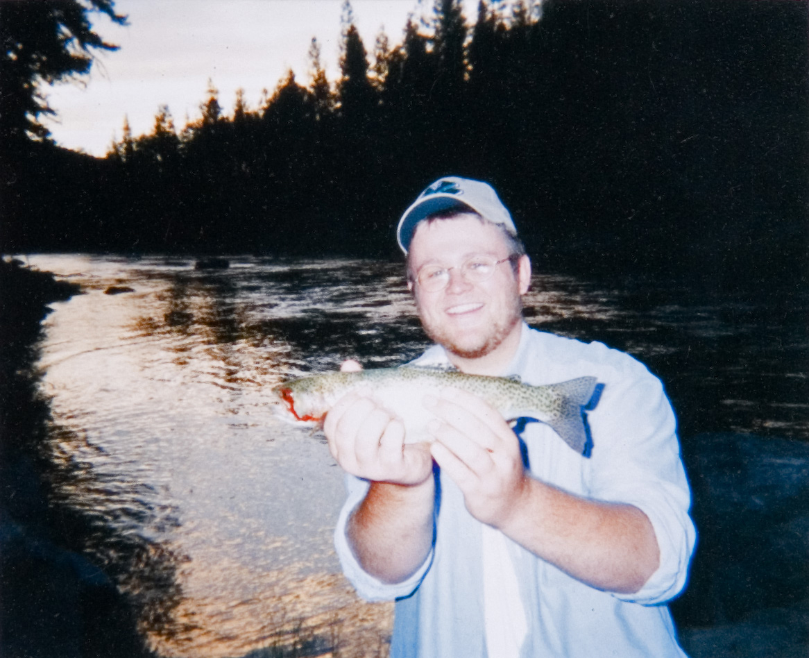 A student in a hat holding up a fish that he caught in a lake.
