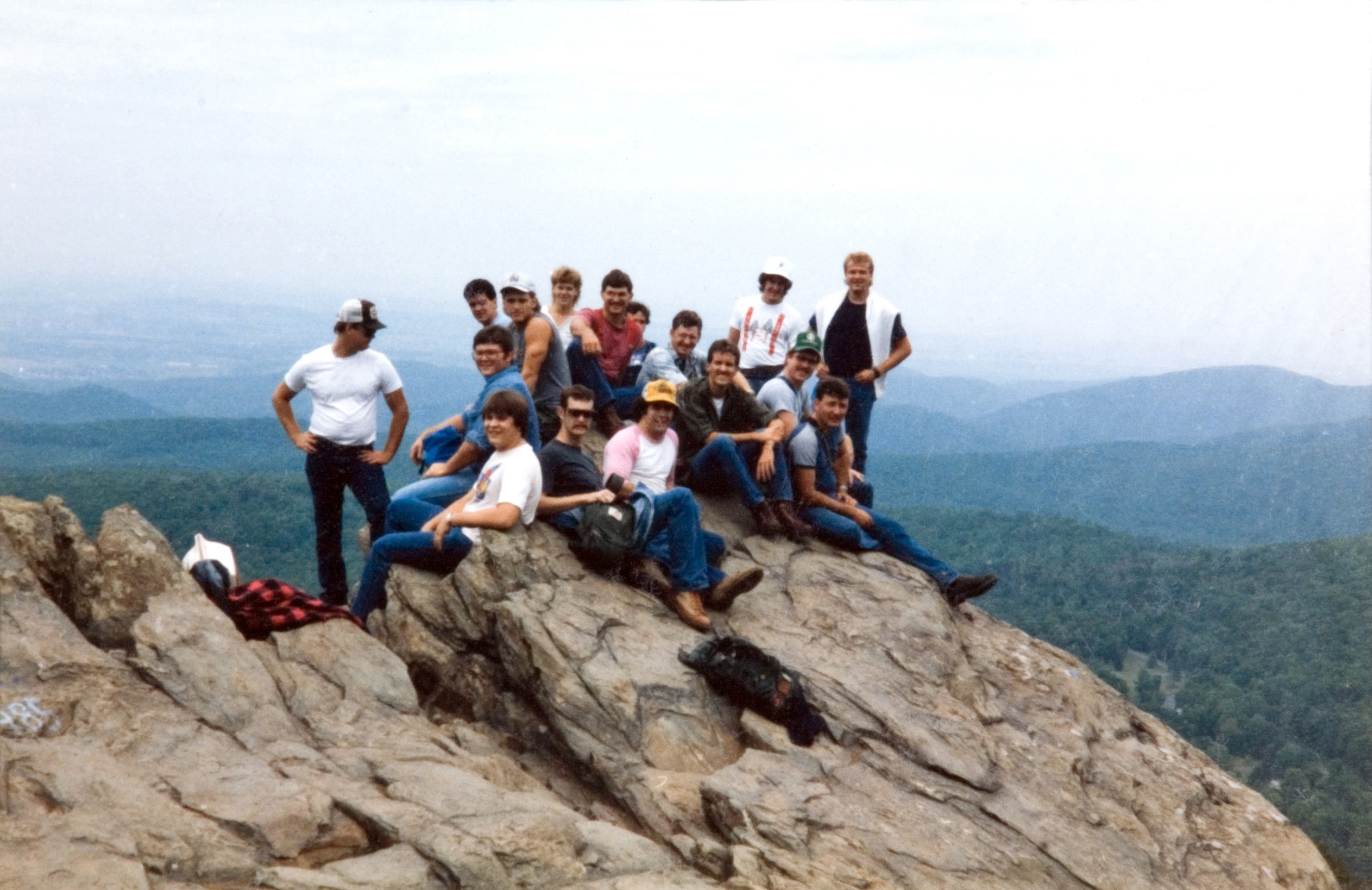A group of 16 students standing and sitting on top of a cliff that overlooks a mountain range.
