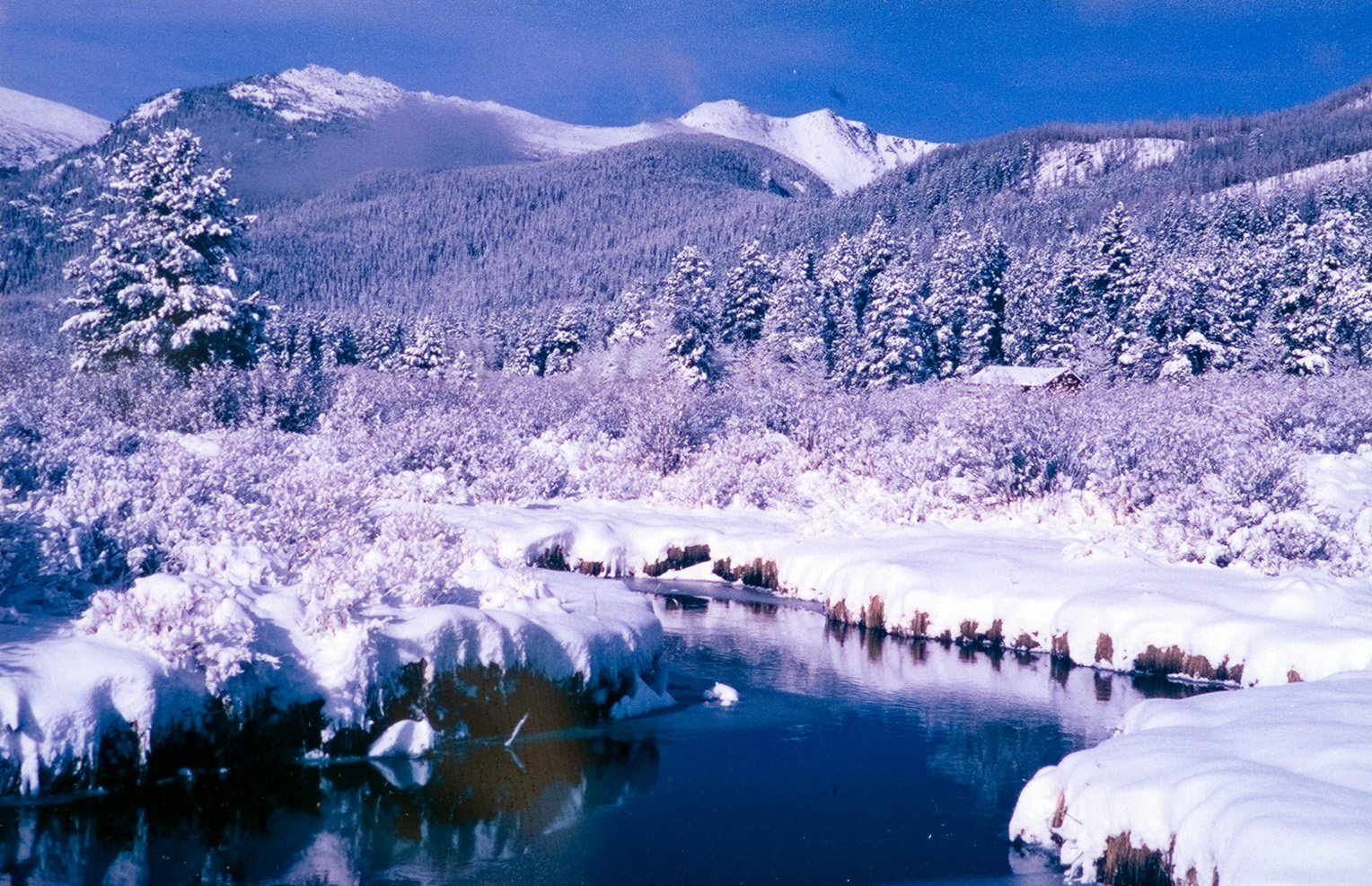 A panoramic view of a still stream next to a snow-covered mountain.