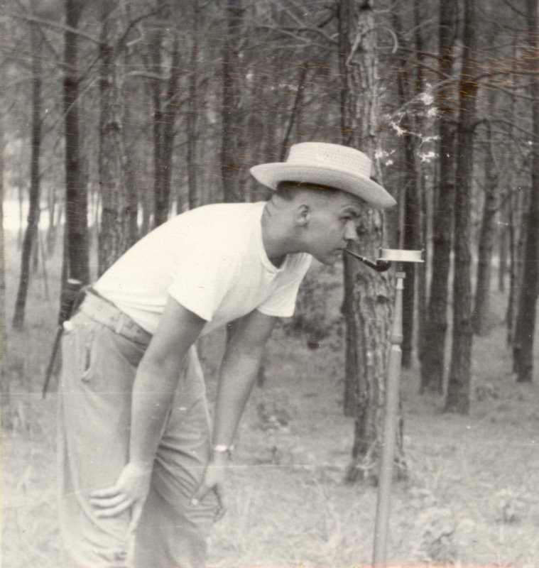 A man with a pipe in his mouth is bent over in the forest looking at equipment.