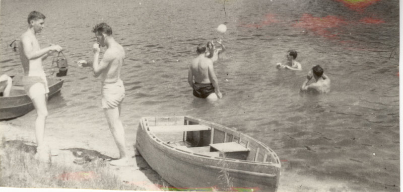 People play in the water while others stand on the shore next to the boats.
