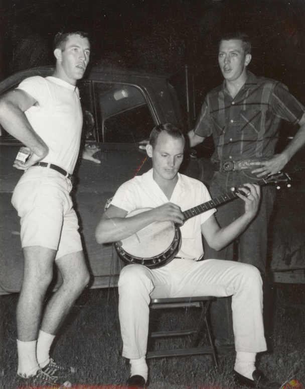 One man is sitting and playing the banjo with two other men standing next to him.