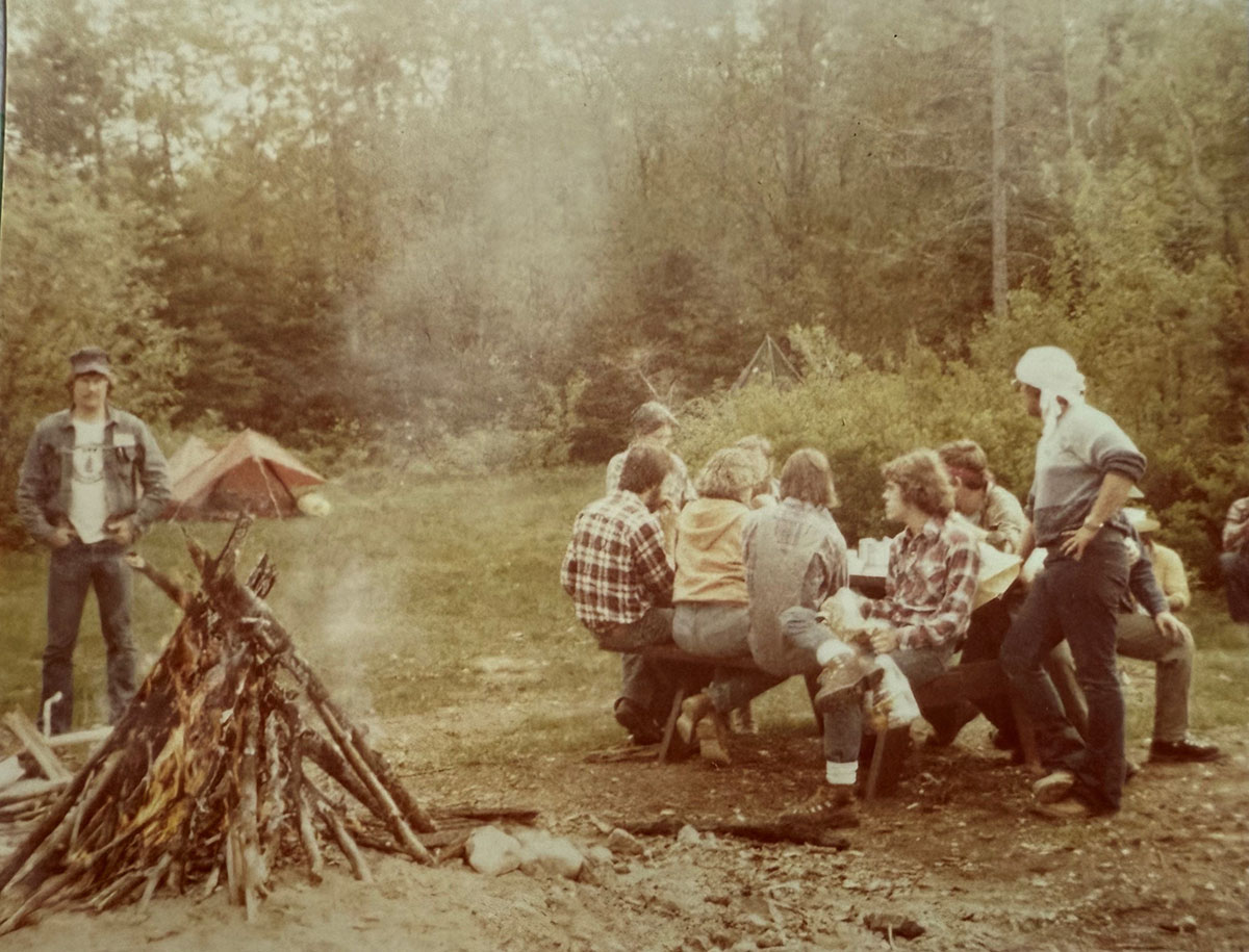 A group of people on the right sitting by a picnic table and a large fire on the left in the woods. A group of people on the right sitting by a picnic table and a large fire on the left in the woods.