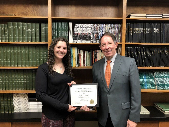 Carly Herndon is accepting her award from Jim Ansley in front of a book shelf. Carly Herndon is accepting her award from Jim Ansley in front of a book shelf.