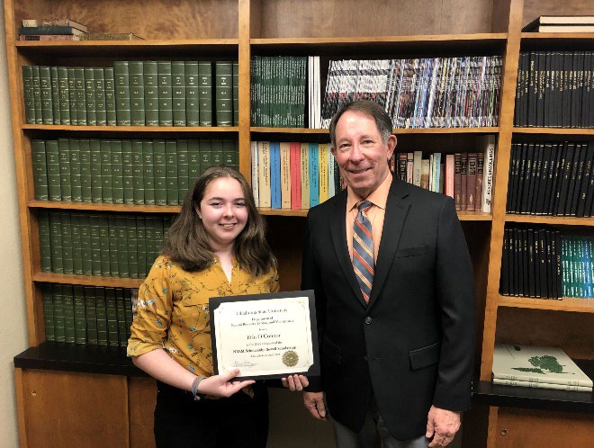 Erin O'Connor is accepting her award from Jim Ansley in front of a book shelf. Erin O'Connor is accepting her award from Jim Ansley in front of a book shelf.