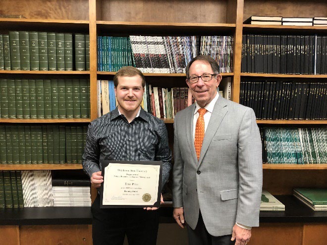 Evan Price is accepting his award from Jim Ansley in front of a book shelf Evan Price is accepting his award from Jim Ansley in front of a book shelf