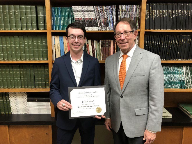 Jackson Russell is accepting his award from Jim Ansley in front of a book shelf Jackson Russell is accepting his award from Jim Ansley in front of a book shelf