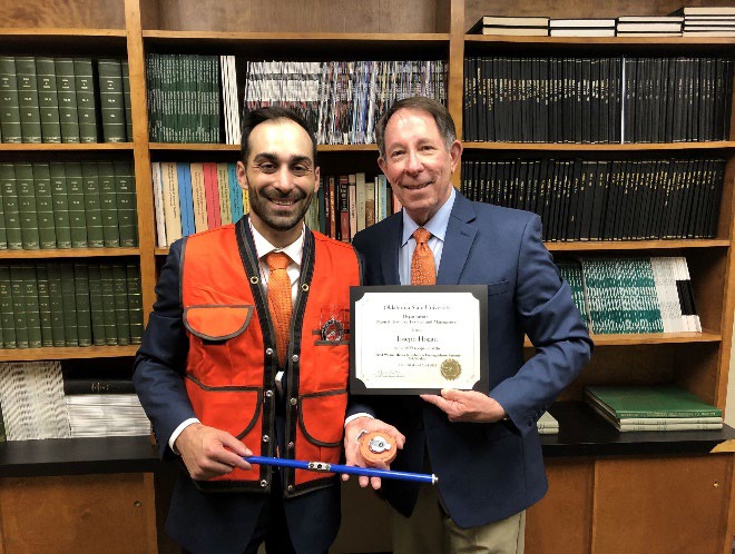 Loseph Hogan is accepting his award with a ornage west on from Jim Ansley in front of a book shelf Loseph Hogan is accepting his award with a ornage west on from Jim Ansley in front of a book shelf