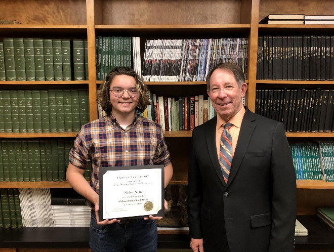 Nathan Stene is accepting his award from Jim Ansley in front of a book shelf. Nathan Stene is accepting his award from Jim Ansley in front of a book shelf.