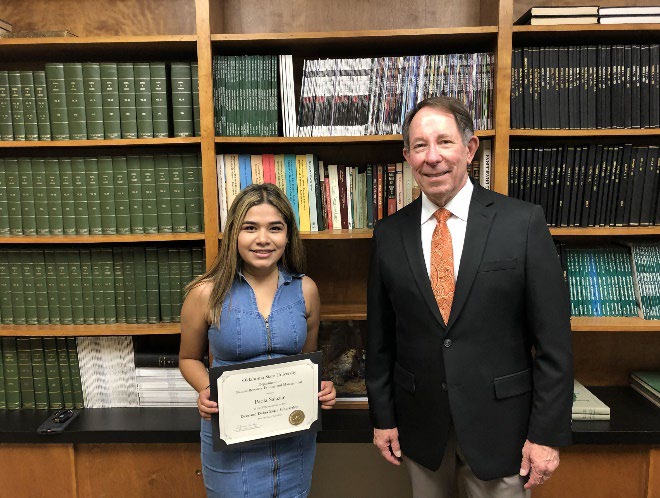 Paola Salazar is accepting her award from Jim Ansley in front of a book shelf. Paola Salazar is accepting her award from Jim Ansley in front of a book shelf.