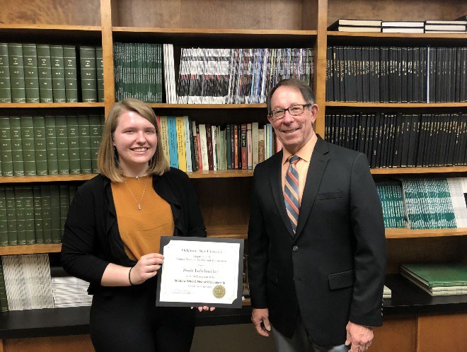 Ronie Loffelmacher is accepting her award from Jim Ansley in front of a book shelf. Ronie Loffelmacher is accepting her award from Jim Ansley in front of a book shelf.