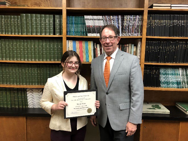 Maddi Watts is accepting her award from Jim Ansley in front of a book shelf. Maddi Watts is accepting her award from Jim Ansley in front of a book shelf.