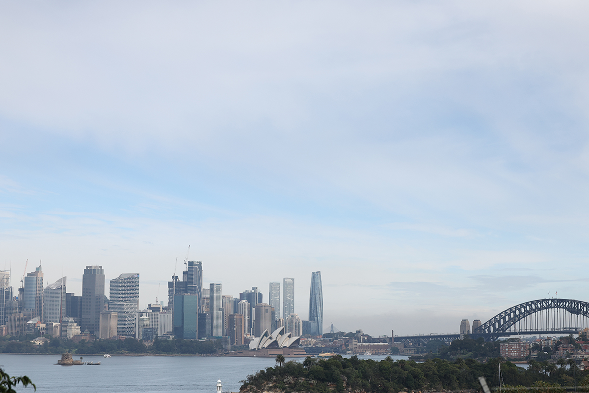 Panoramic view of Sydney Harbour featuring the Sydney skyline across the water. The Sydney Opera House sits near the center of the image, with the Sydney Harbour Bridge visible on the right. Modern high-rise buildings line the waterfront, while a tree-covered foreground and calm harbor waters frame the scene beneath a partly cloudy blue sky.